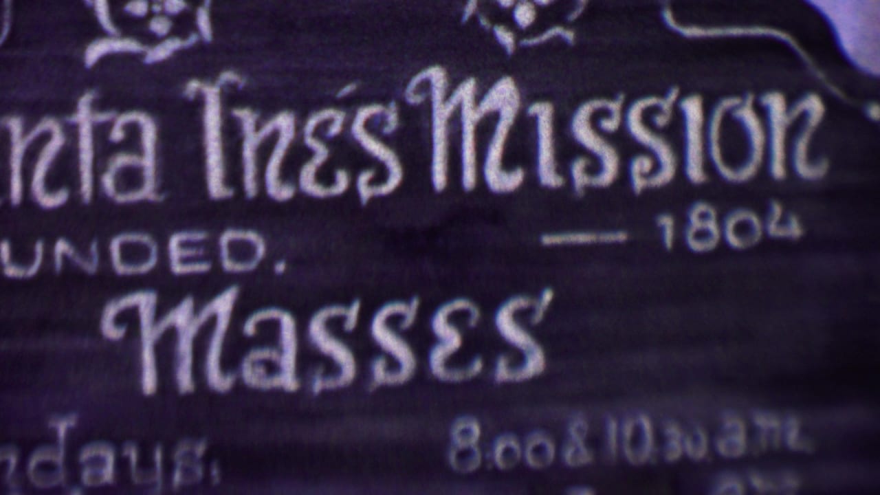 Close-up from 1955 Solvang footage showing white lettering on a dark Santa Ines Mission sign with mass-times text partially visible.