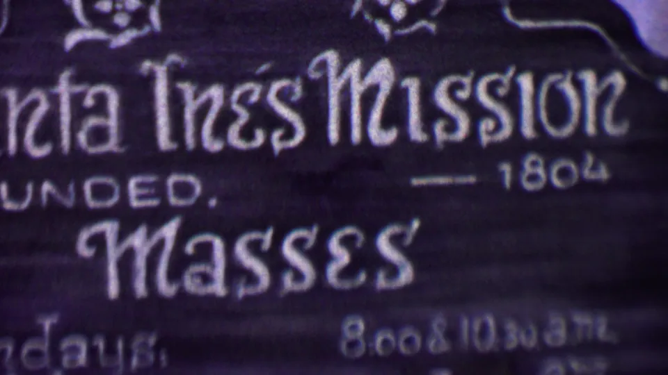 Close-up from 1955 Solvang footage showing white lettering on a dark Santa Ines Mission sign with mass-times text partially visible.