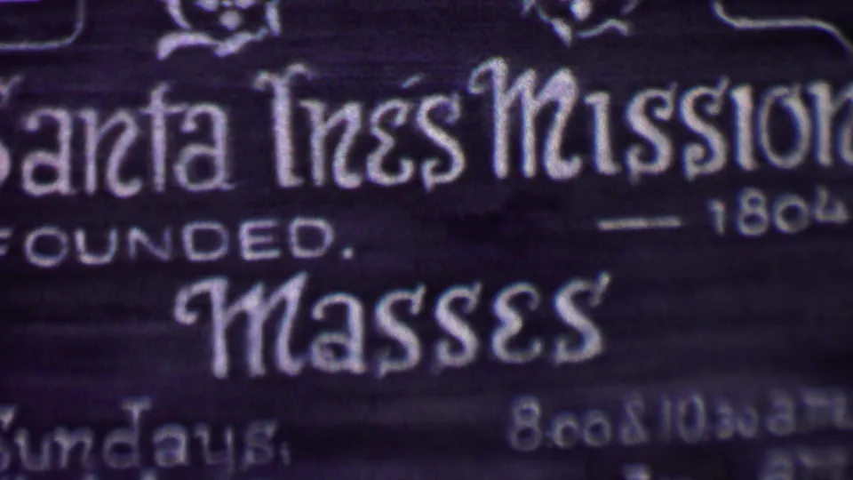Close framing of the Santa Ines Mission sign in Solvang, with founded-1804 text and mass information visible in soft 8mm focus.