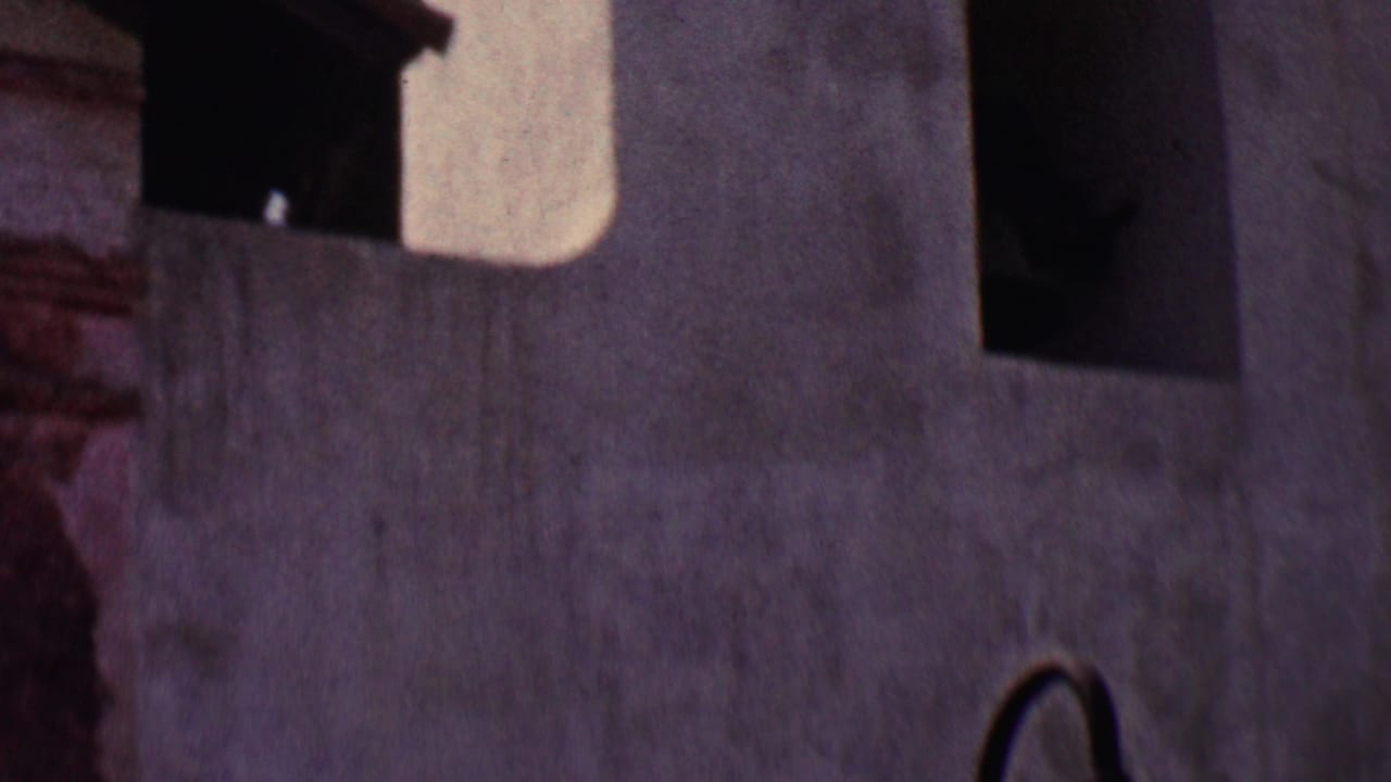 1955 Solvang home-movie frame showing a close low-angle view of white stucco building walls, shadowed arched openings, and dark roof trim.