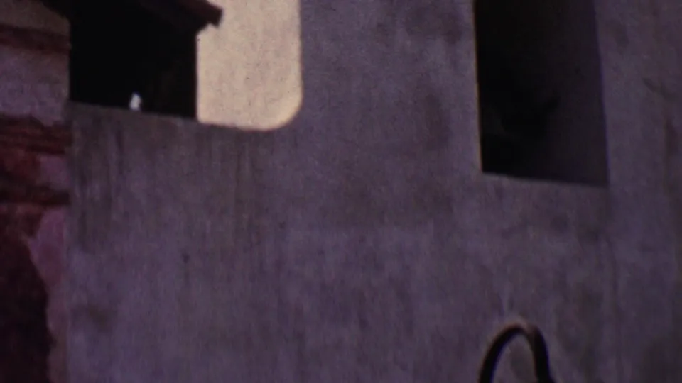 1955 Solvang home-movie frame showing a close low-angle view of white stucco building walls, shadowed arched openings, and dark roof trim.
