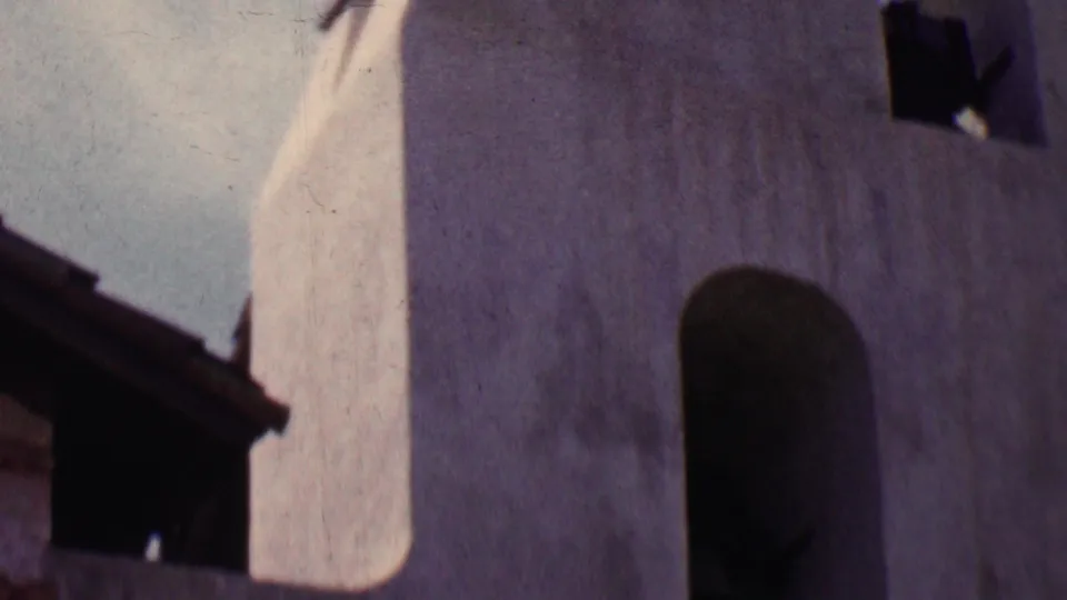 1955 Solvang frame with the camera angled up toward a white stucco bell-tower section, shadowed arch, roofline, and a small dark bell or bell housing.