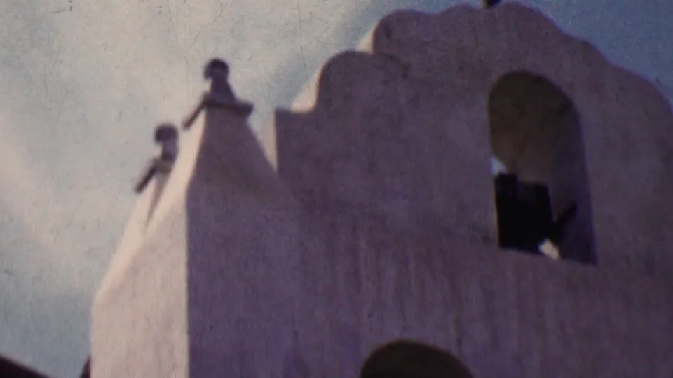 1955 Solvang home-movie frame showing a white stucco bell tower with curved parapet edges, two small finials, an arched opening, and pale sky behind it.