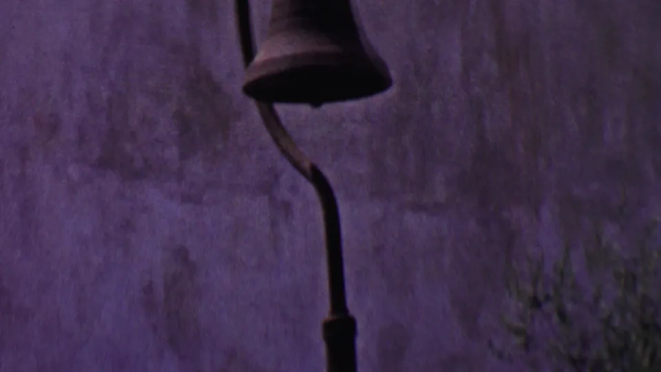 Solvang, California 1955 frame showing a hanging bell mounted on a curved bracket against a textured stucco exterior with sparse greenery below.
