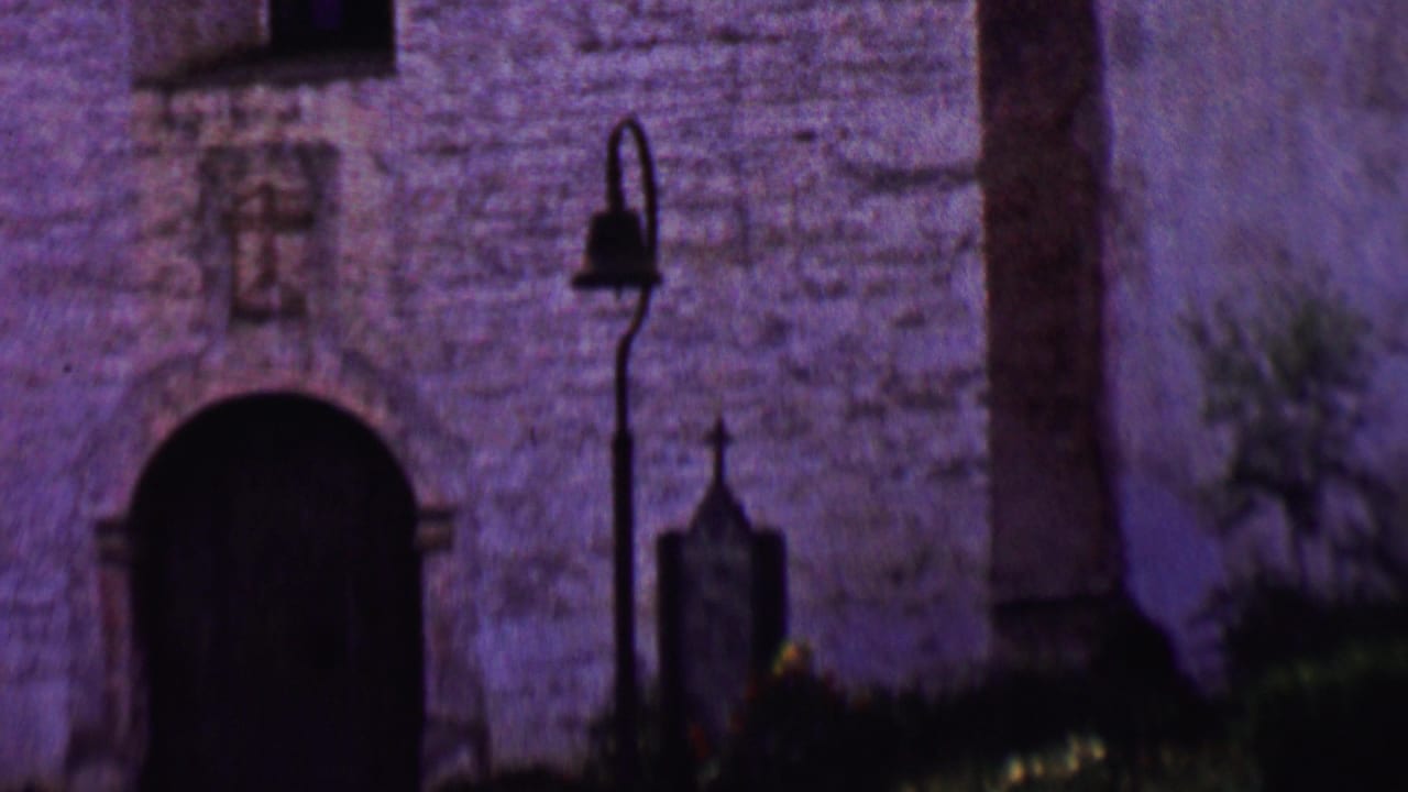 Arched doorway in a stone church wall with a bell post and cross marker nearby.