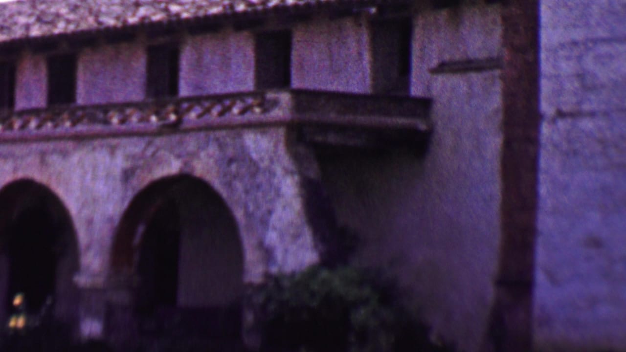 1955 Solvang home-movie frame showing a Spanish-style stucco facade with arched doorways, dark upper windows, tiled roof edging, and a small projecting balcony.