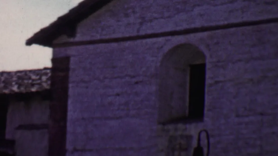 1955 Solvang home-movie frame showing a broad stucco wall with two arched window openings, a sloped roof edge, and part of a lower adjoining structure.