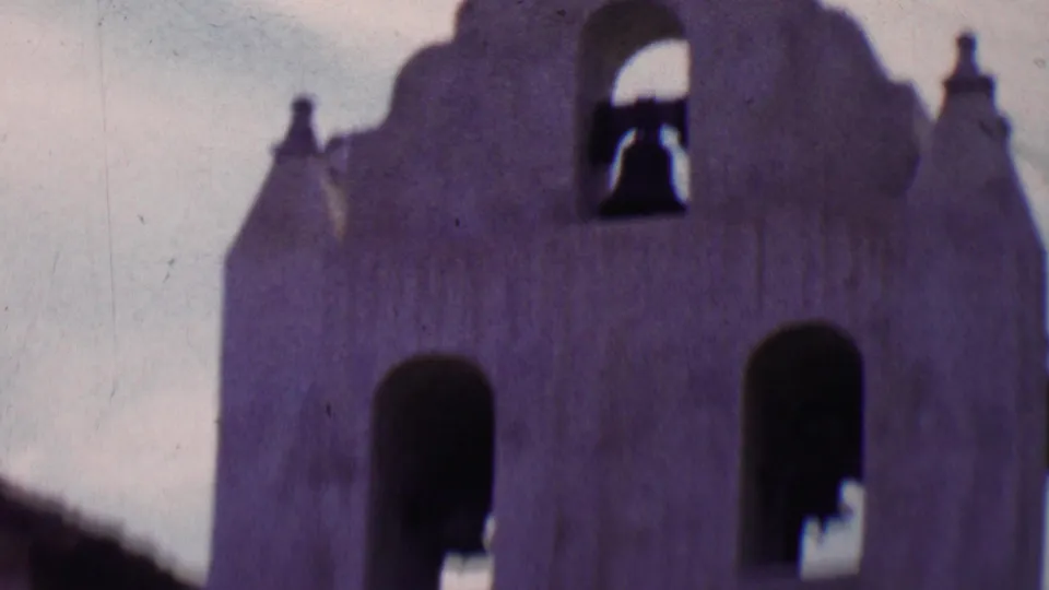 1955 Solvang 8mm view of a church or mission-style bell tower facade, with arched openings, visible bells, stucco forms, and pale daytime sky.