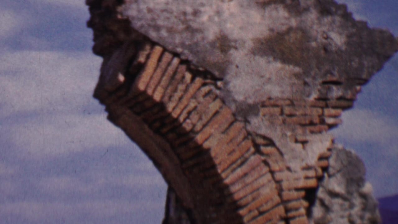 1955 Solvang-area 8mm frame showing the upper portion of a weathered brick and stone mission arch ruin with blue sky and hills behind it.