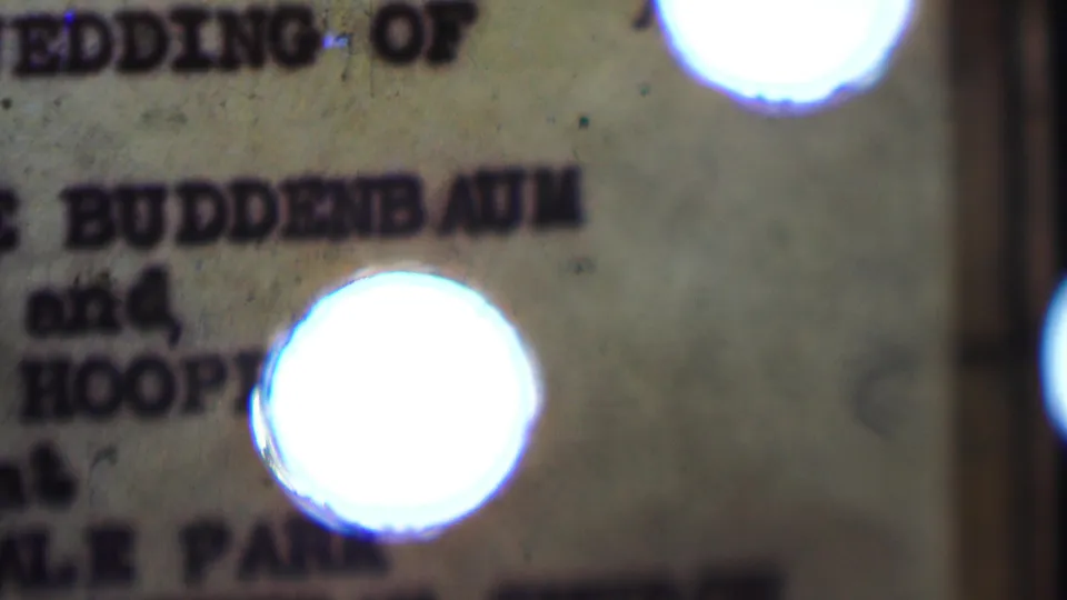 Soft closing frame of the typewritten wedding invitation, with Buddenbaum and Hooper partly visible and a bright circular flare over the paper surface.