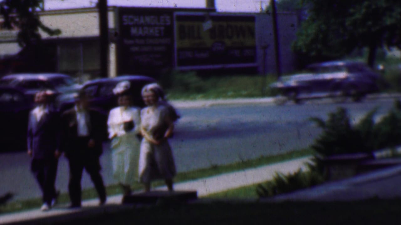 In Detroit, Michigan circa 1950, four adults in suits, dresses, and hats walk along a sunny sidewalk with classic cars and storefront signage in the background.