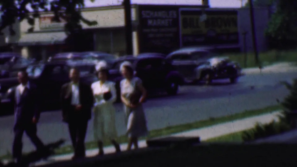 Adults in mid-century clothing walk near a dark sedan on a Detroit sidewalk, with moving and parked cars and storefront signs visible across the street.