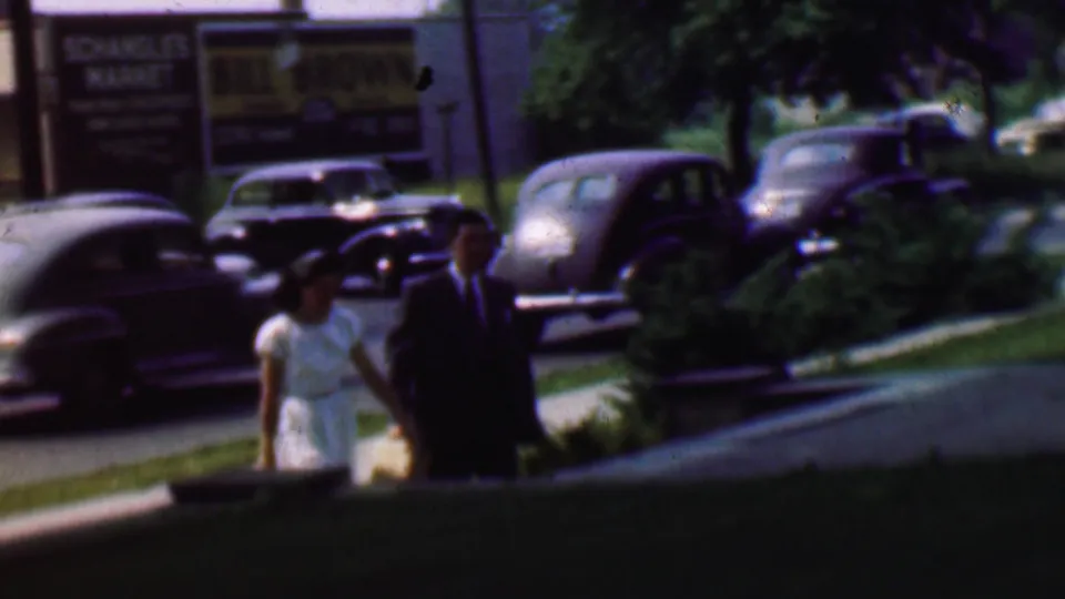 A woman in a light dress and a suited man walk along a Detroit sidewalk near rounded mid-century parked cars and a visible commercial sign.
