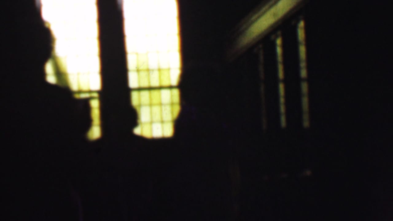In a 1950 Detroit church interior, dark silhouetted figures stand or move near tall warm-lit grid-patterned windows, with railings or stair lines barely visible.