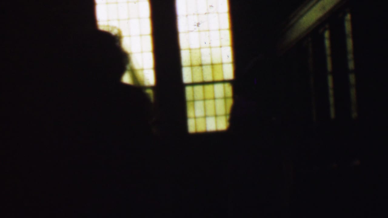 A large dark figure shape crosses in front of bright window panels and railings.