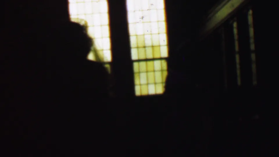 A silhouetted person appears in the foreground of the dim church interior, partly blocking warm rectangular window light and faint stair or balcony railings.