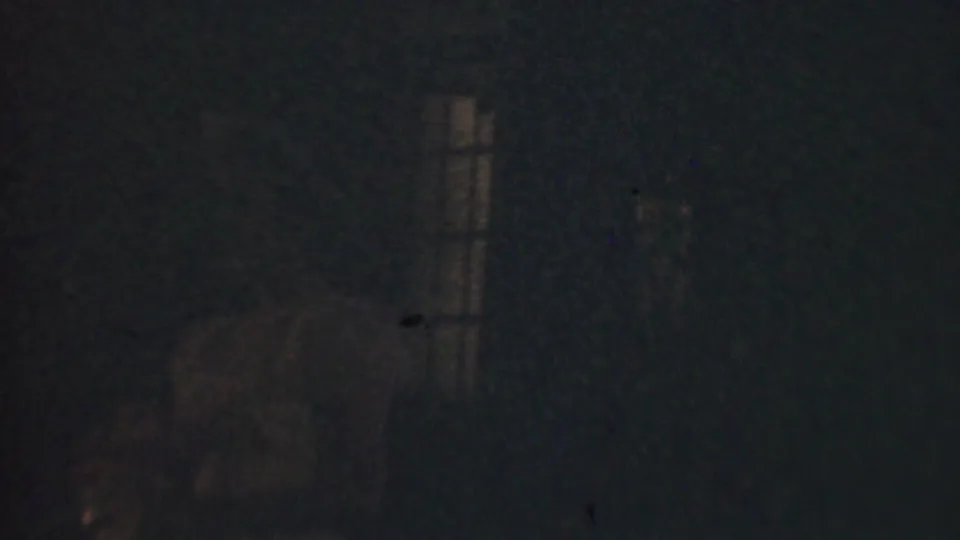 Extremely dark 1950 Detroit 8mm frame with a barely visible woman near a dim vertical window light and heavy film grain.
