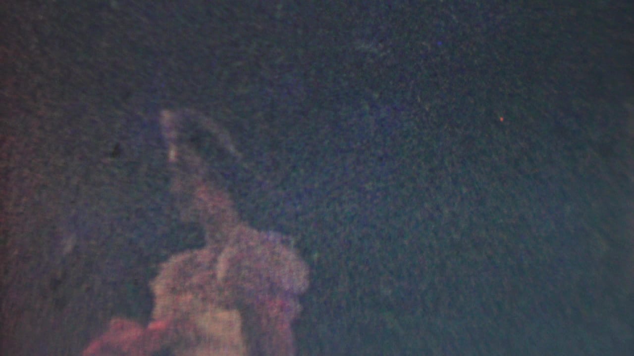 Dim frame with a woman in a pale dress partly visible against darkness.