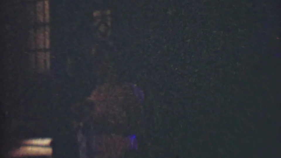 Grainy 1950 Detroit 8mm frame showing a blurred figure low in the frame, a bright rectangular light, and a mostly dark background.