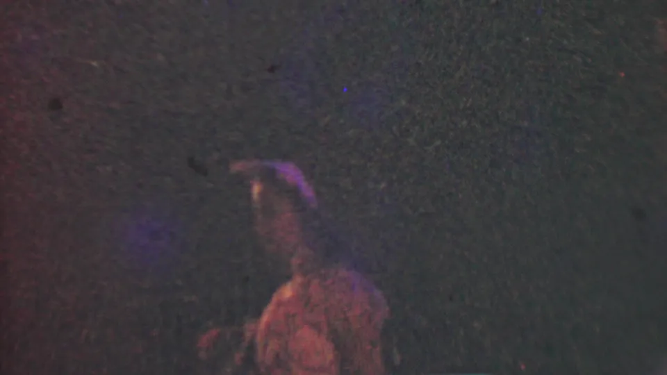 In 1950 Detroit 8mm footage, a young woman wearing a dress and hair bow appears dimly at night amid reddish-purple light and heavy film grain.
