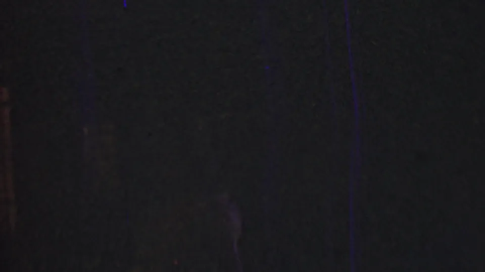 A very underexposed 1950 Detroit home-movie frame shows faint vertical forms, shadowed background detail, and colored film artifacts with little clearly identifiable action.