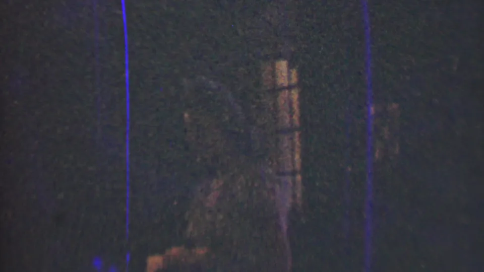 The young woman appears faintly near vertical window-like lines in the dark 1950 Detroit 8mm frame, with her dress barely visible through grain and scratches.