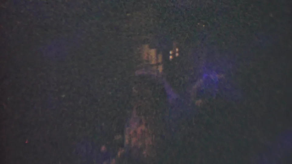 Low-light 1950 Detroit home-movie frame showing indistinct people in a dark room with small warm light points and visible film scratches.