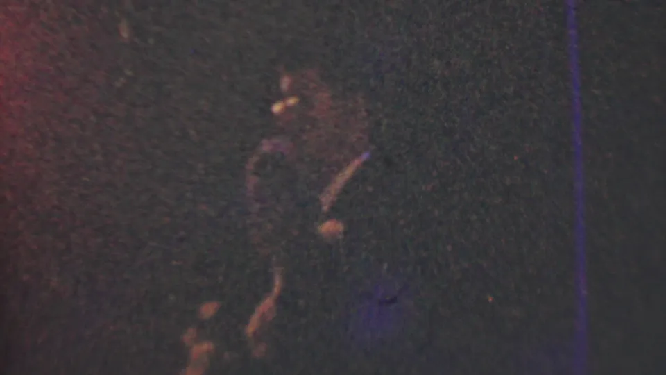 Underexposed 1950 Detroit home-movie frame with a partially visible figure at left, heavy grain, and blue-purple scratch marks across the image.