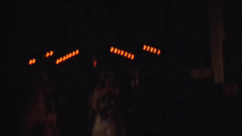 Dark 1950 Detroit home-movie frame with rows of warm orange lights, indistinct people below, and soft grainy exposure in an indoor celebration setting.