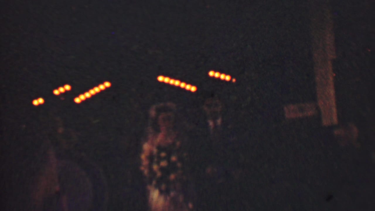 In supported 1950 Detroit home-movie footage, a bride in a veil and a groom in a dark suit walk through a dim outdoor wedding scene with guests and string lights behind them.