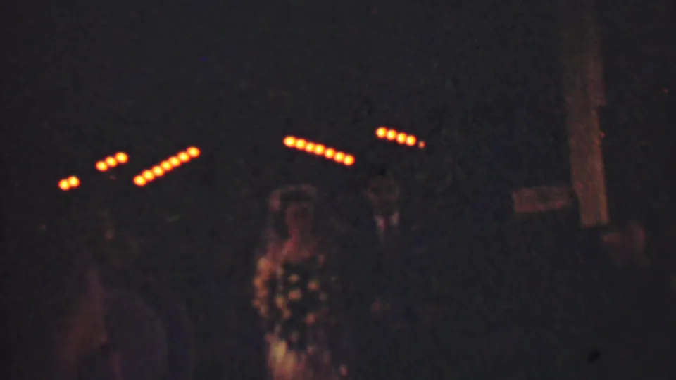 In supported 1950 Detroit home-movie footage, a bride in a veil and a groom in a dark suit walk through a dim outdoor wedding scene with guests and string lights behind them.