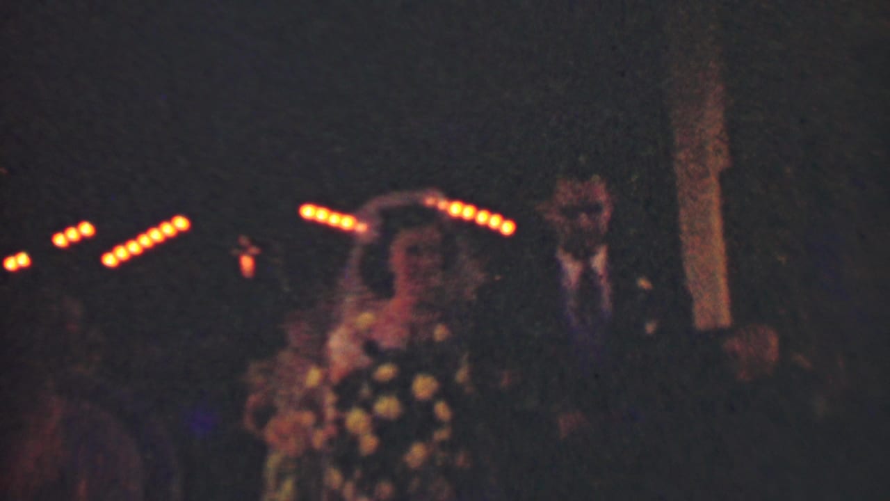 Bride holding flowers stands or walks beside groom in low light with guests and string lights in the background.