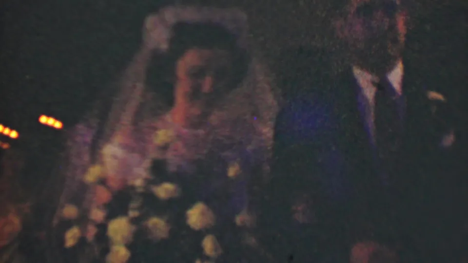 A closer 1950 Detroit wedding frame shows the bride's veil and bouquet beside the groom's dark suit, with dim guests and warm lights receding behind them.