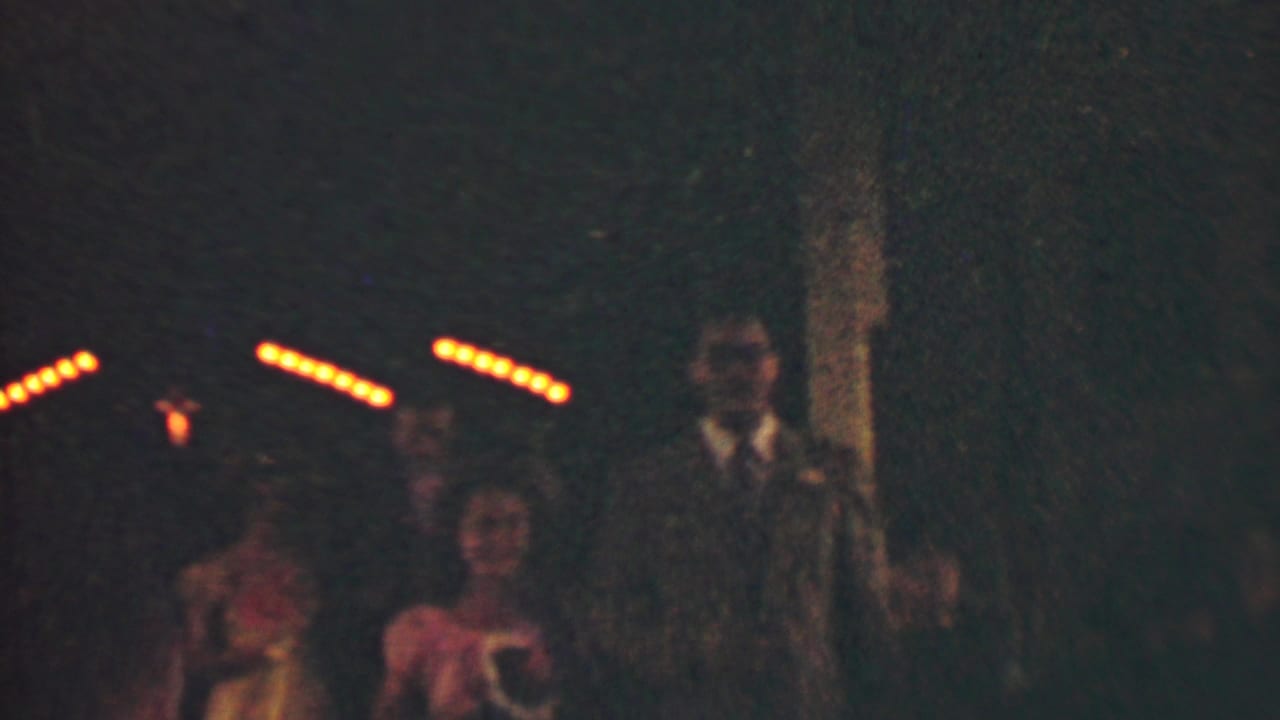 In 1950 Detroit home-movie footage, women in dresses and men in suits appear to move through a dark formal-event aisle under warm streaks of light.