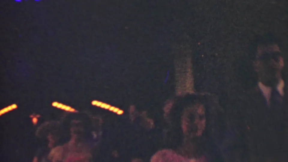 A suited man stands or walks near women carrying or wearing flowers in a dark 1950 Detroit formal-event scene, with orange lights crossing the background.