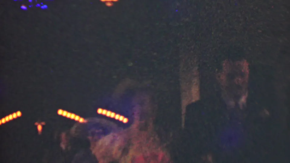 Blurred formal-event guests or participants pass the handheld 8mm camera in a dim Detroit setting, with a suited man partly visible behind the motion.