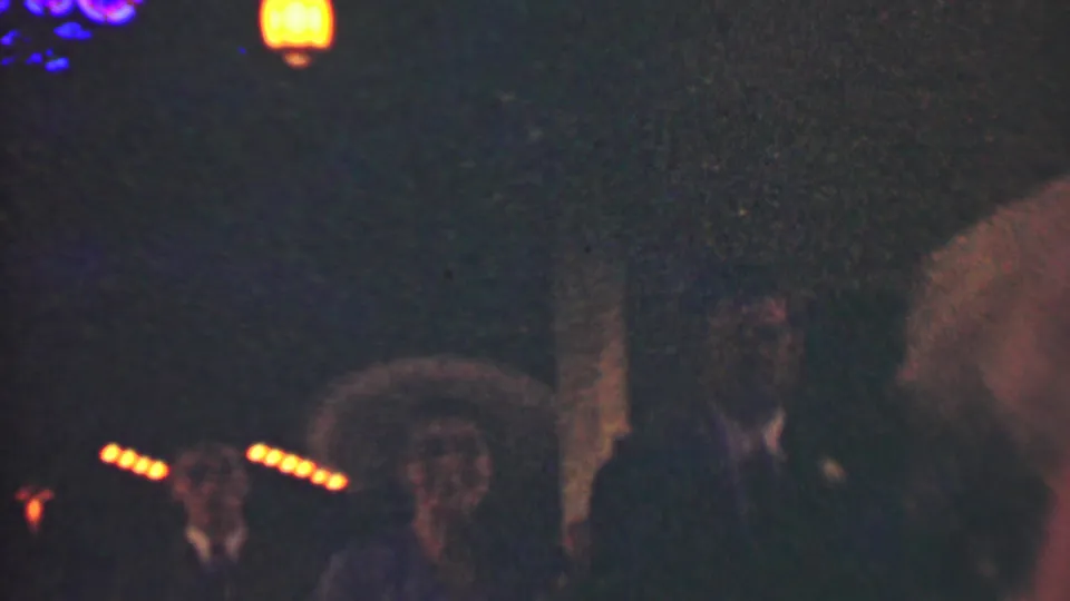 A woman wearing a hat or headpiece appears beside suited men in the dim 1950 Detroit home-movie scene, with the procession continuing through warm background lights.