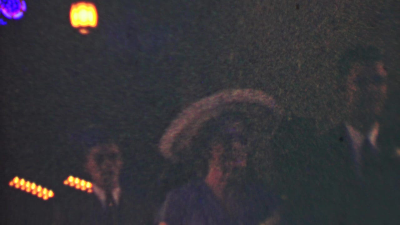 Dim 1950 Detroit home-movie frame showing formally dressed adults at night beneath warm string lights and lantern-like glows.