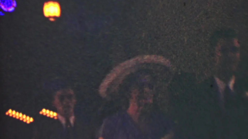 Dim 1950 Detroit home-movie frame showing formally dressed adults at night beneath warm string lights and lantern-like glows.
