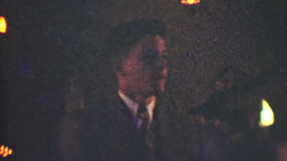 Low-light 1950 Detroit frame with a man in a dark suit and tie visible among other formal guests and warm event lights.