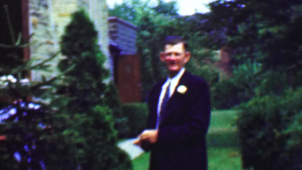 Daytime 1950 Detroit home-movie frame showing a man in a dark suit and tie standing in a manicured garden with a stone building behind him.
