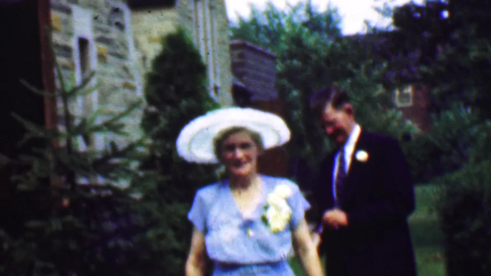 Daytime 1950 Detroit frame showing a smiling couple outdoors, with the woman in a light blue dress and broad white hat beside a man in a dark suit and boutonniere.