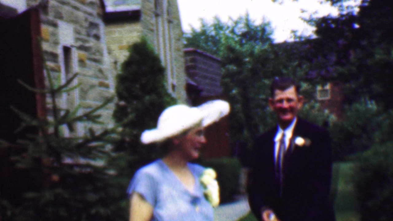 In a 1950 Detroit home-movie frame, a woman in a pale blue formal dress and wide white hat stands outdoors near a man in a dark suit beside stone buildings and greenery.