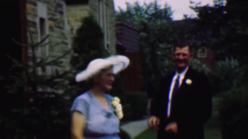 A 1950 Detroit wedding home-movie frame shows a woman in a pale blue dress and white hat walking near a man in a dark suit, with shrubs and stone buildings behind them.