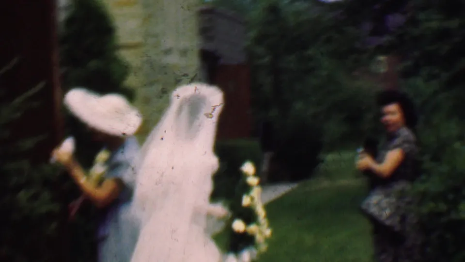 This 1950 Detroit home-movie frame shows a veiled bride and formally dressed guests moving across a lawn, with floral arrangements and garden shrubs in view.