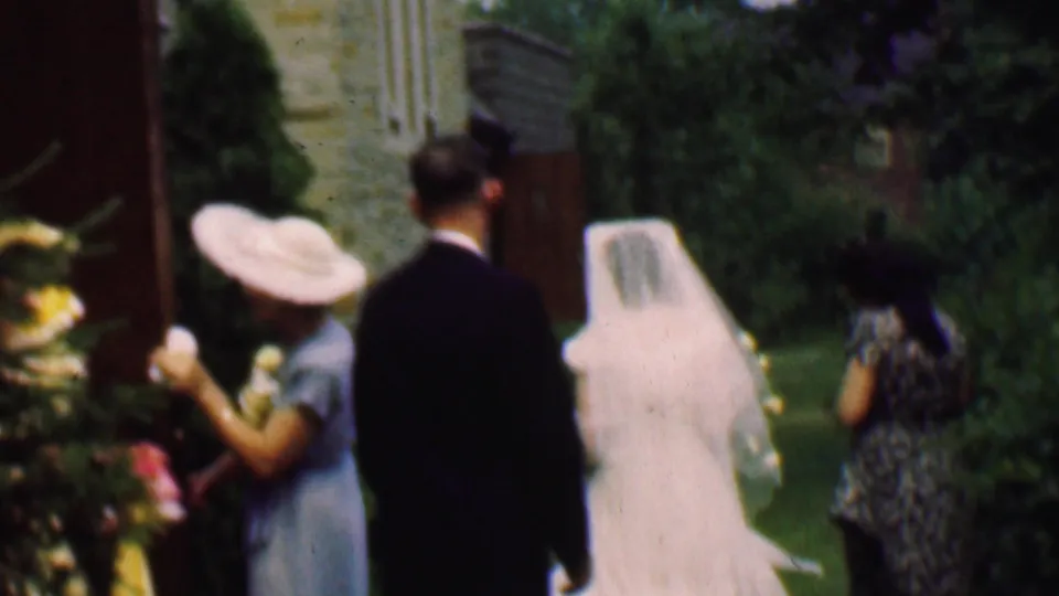 A 1950 Detroit wedding home-movie frame shows the veiled bride, a man in a dark suit, and women in formal dresses walking through a garden with stone buildings and greenery.