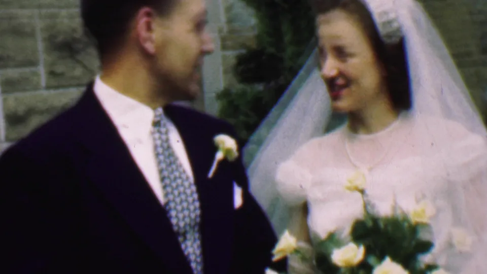 The bride and groom share a candid smile outside the church in this 1950 Detroit home-movie frame, with the bride holding a bouquet of pale yellow flowers.