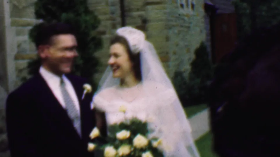 The 1950 Detroit bride and groom stand close together outside the stone church, smiling with the bouquet centered while a dark foreground figure partially enters the frame.