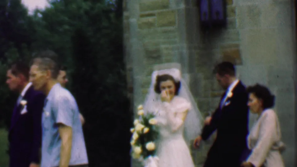 The bride stands with her bouquet near the church entrance while the groom and several guests move through the frame in this 1950 Detroit wedding home movie.