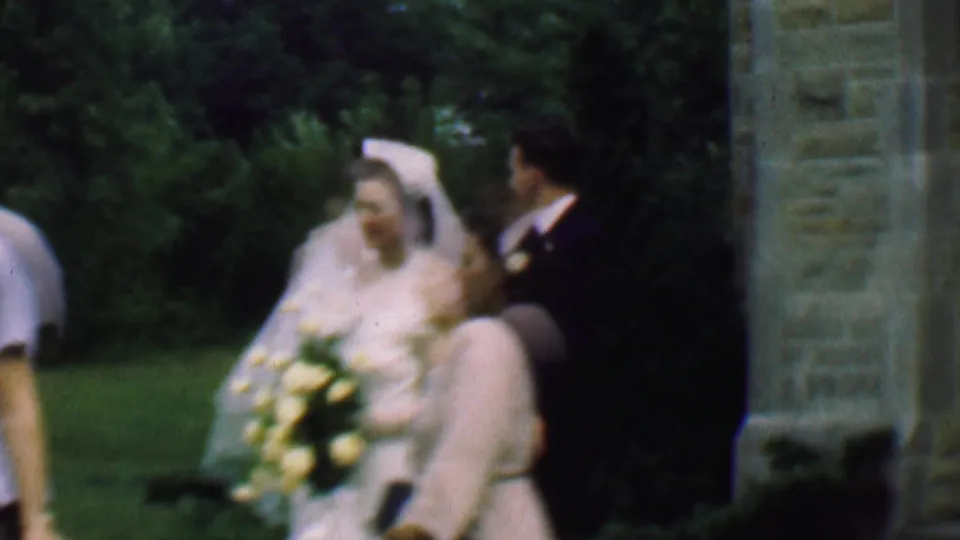 The bride and groom pause near adult guests by the stone church exterior and greenery, with the bride's veil and bouquet prominent in the frame.